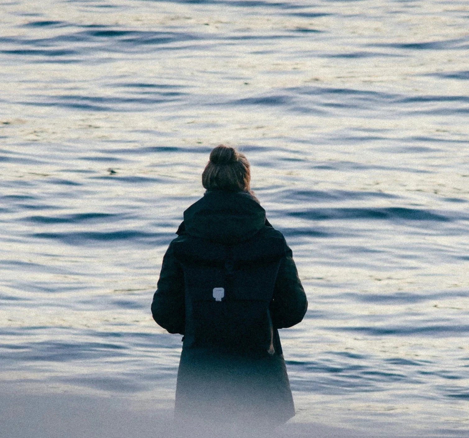 En person med mörkt hår i en knut, sedd bakifrån, står vid vattnet och ser ut mot havet eller sjön under en molnig dag, iklädd svart jacka.
