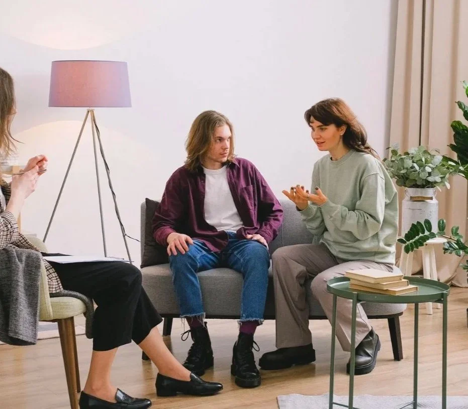 Två kvinnor och en man i ett samtal i ett vardagsrum, med växter och böcker i bakgrunden.