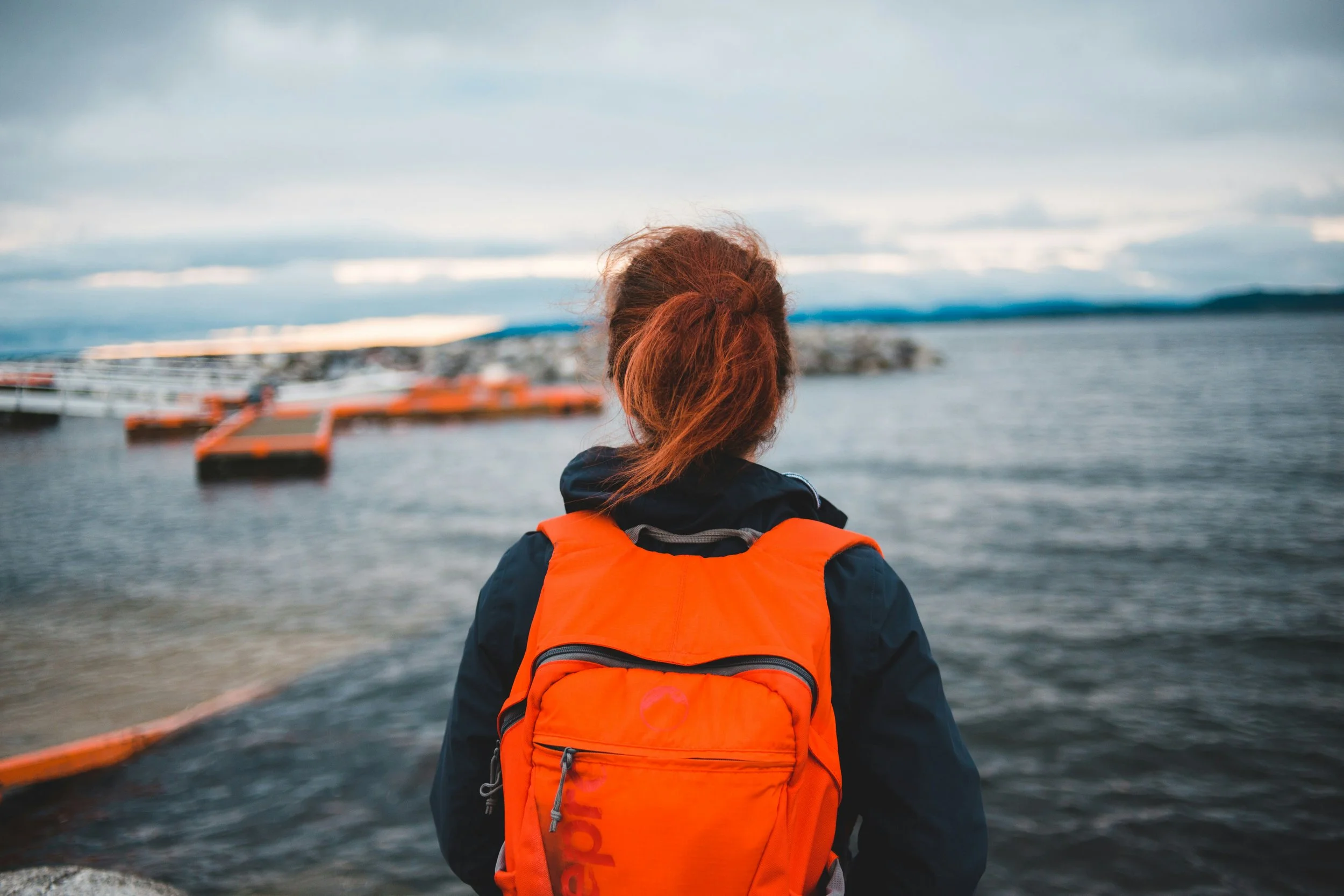 En person med röd hår, bär en orange flytväst, tittar ut över vatten och ett piratskepp i en hamn, molnigt väder.