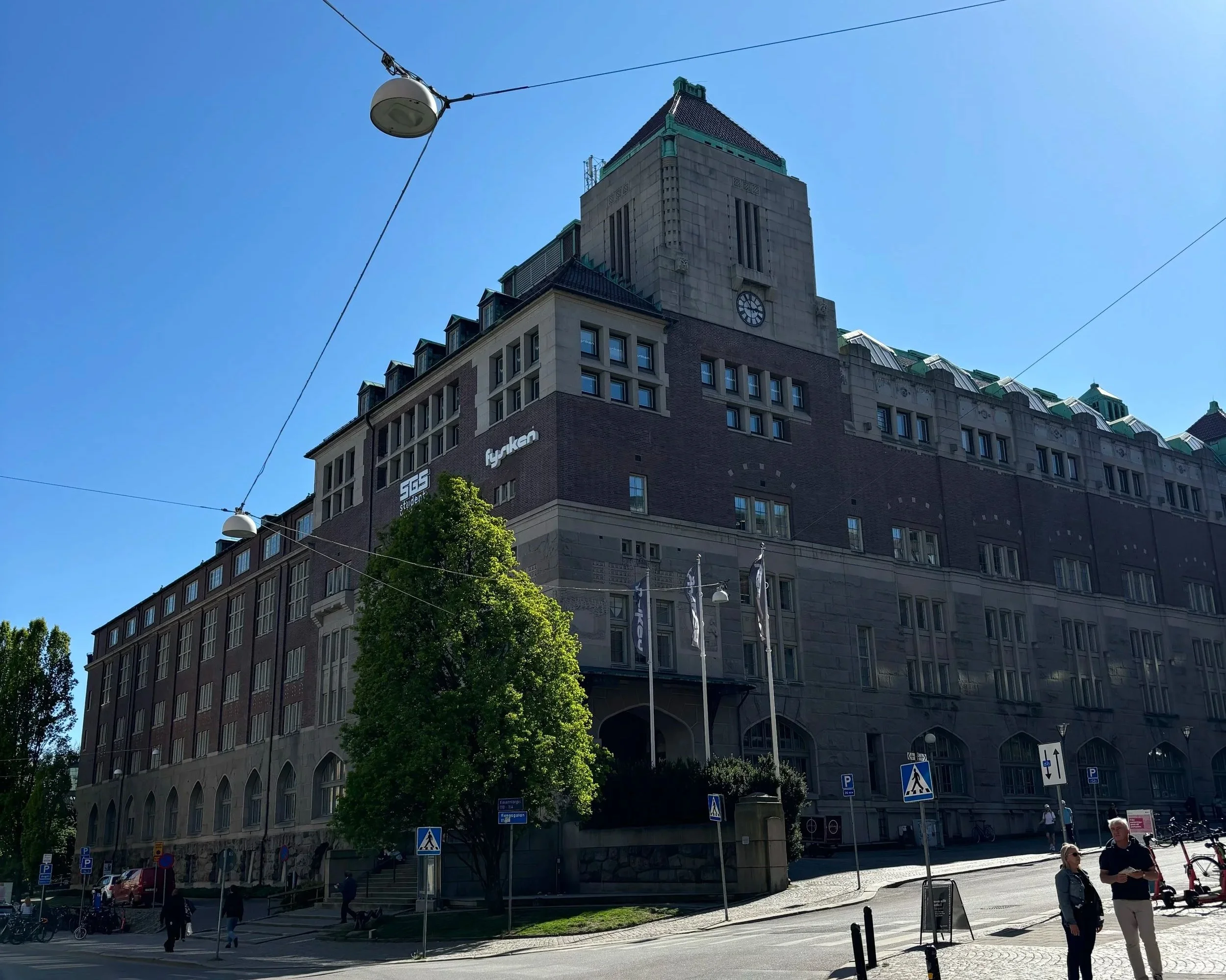 En stadsvy med ett högt byggnadshus som har en klocka och grön takdetalj, omgivet av andra byggnader och tydliga trafikmärken samt människor på gatan under en klar blå himmel.