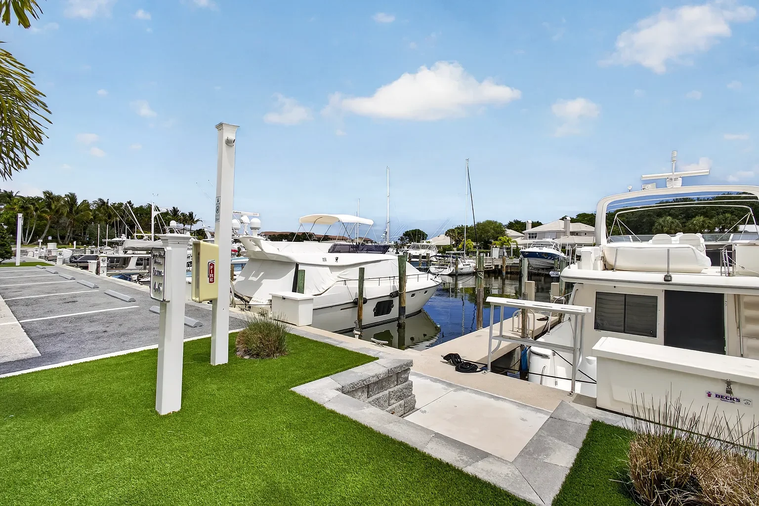 A marina with multiple boats docked along the wooden piers, a parking lot with marked spaces, and a grassy area with small plants, under a partly cloudy sky.