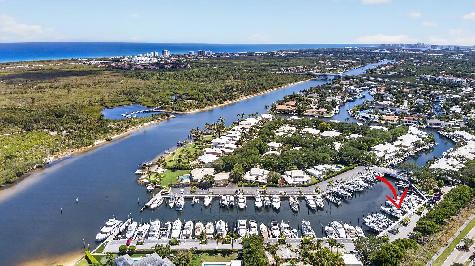 Aerial view of a marina with boats docked along a canal, residential houses surrounding the water, and a distant coastline with high-rise buildings on the horizon.