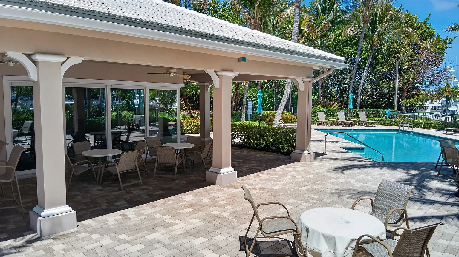 Poolside patio with round table, chairs, and a shaded area, overlooking a swimming pool surrounded by lounge chairs and greenery.