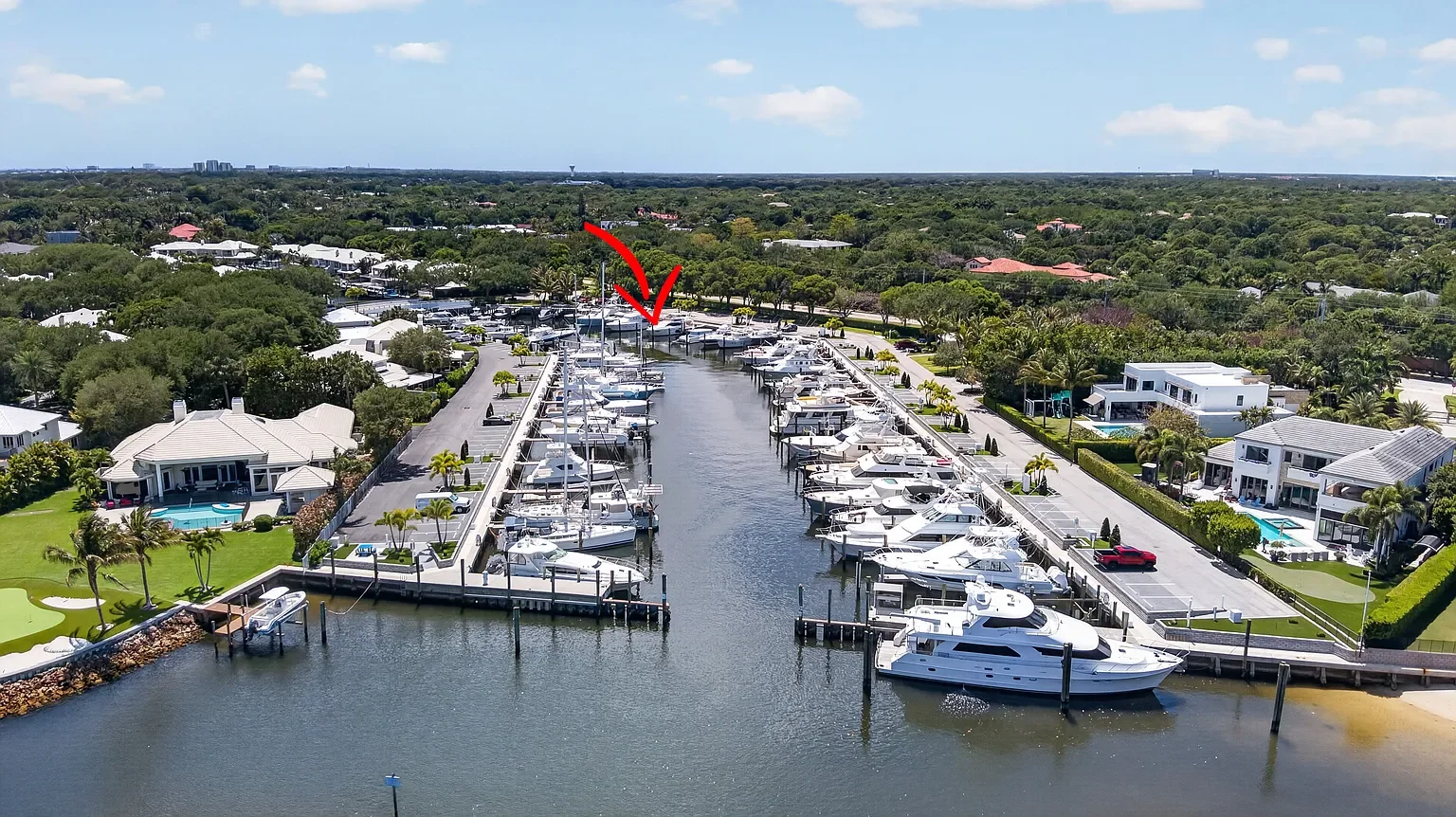 Aerial view of a marina with numerous yachts docked, surrounded by residential houses, some with swimming pools, and greenery, under a partly cloudy sky.
