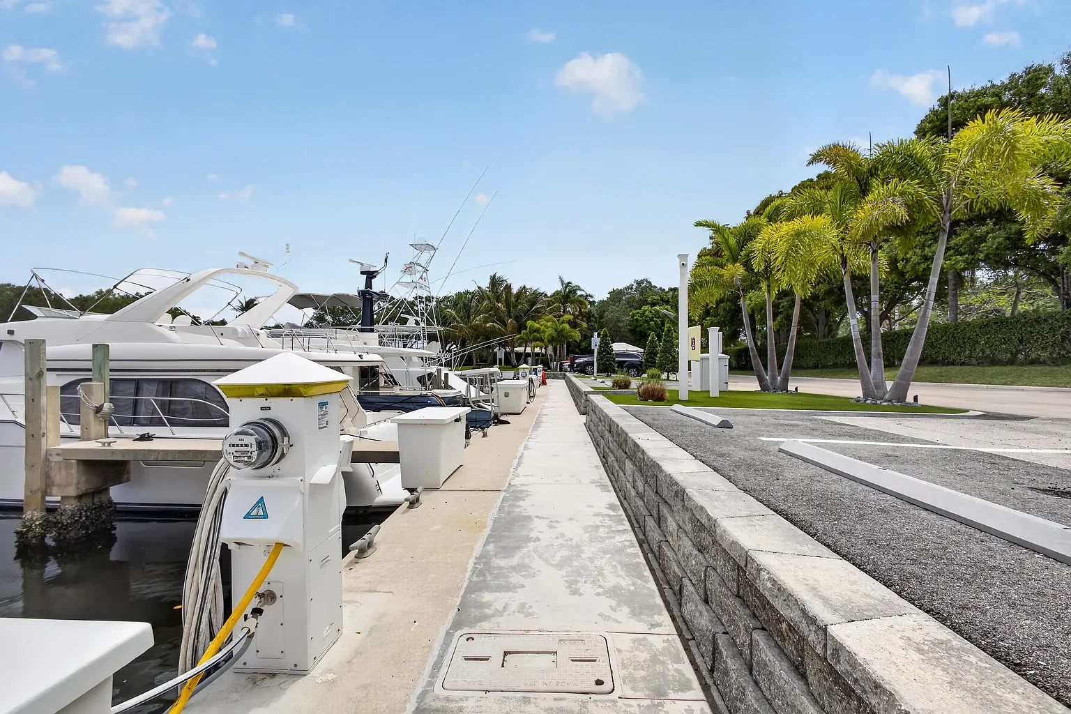 Boats docked at a marina with electric charging stations on a sunny day, palm trees, and a parking lot.