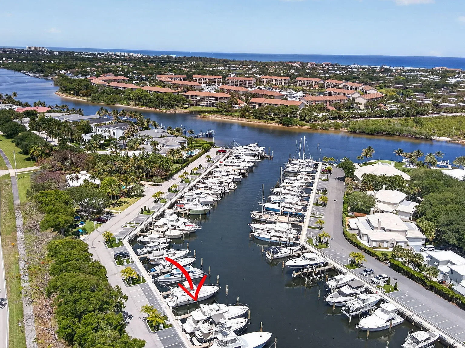 Aerial view of a marina with boats docked, residential buildings, waterway, lush greenery, and distant ocean shoreline under a blue sky.