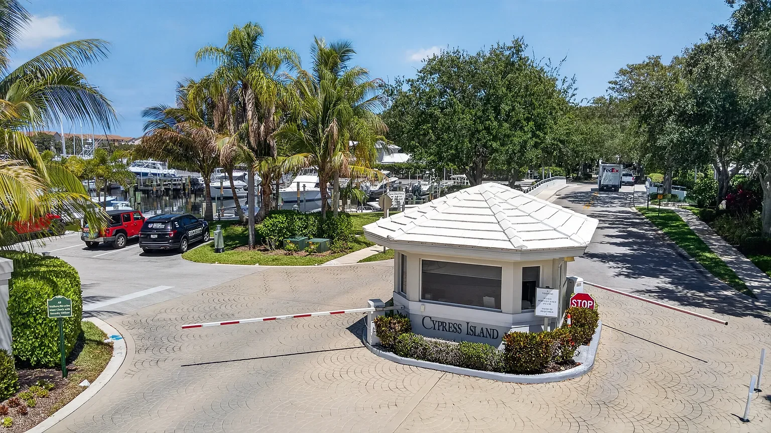 Entrance gate to Cypress Island with guard booth, stop sign, security sign, palm trees, parked cars, marina, and boats in the background.