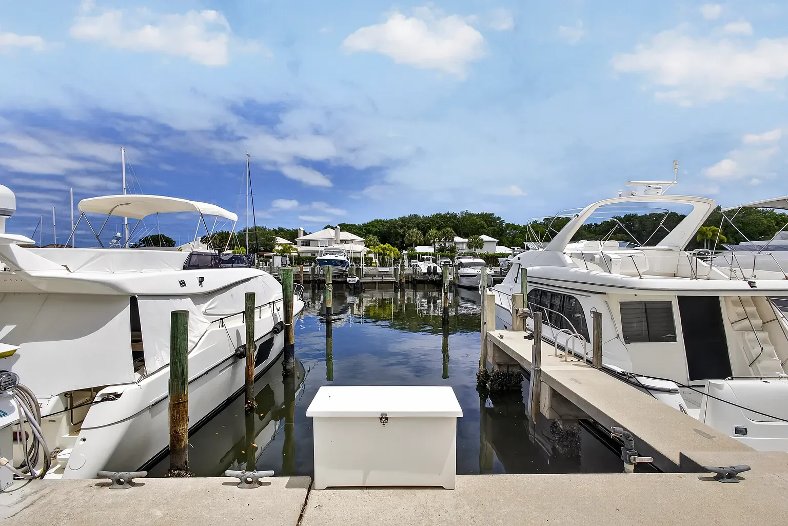 Marina with boats docked at the pier, clear blue sky with some clouds, and houses in the background.
