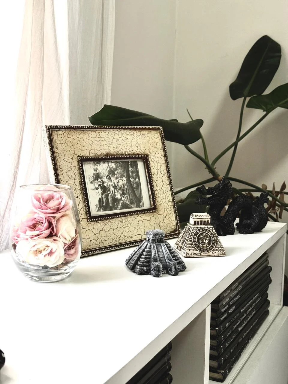 Vaso con rosas de color rosa y blanco, marco de fotos con diseño envejecido, figura de pirámide maya y dos figuras de elefantes de color negro sobre una repisa blanca decorada con libros apilados y una planta de hojas verdes en el fondo.