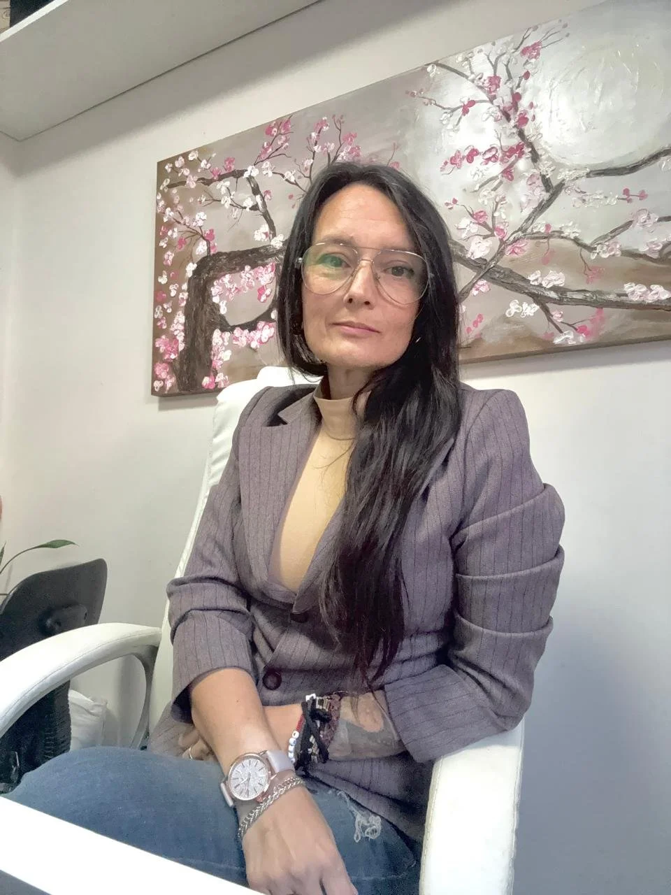Mujer con gafas y cabello largo, sentada en una silla blanca, con un cuadro de flores de cerezo en el fondo.