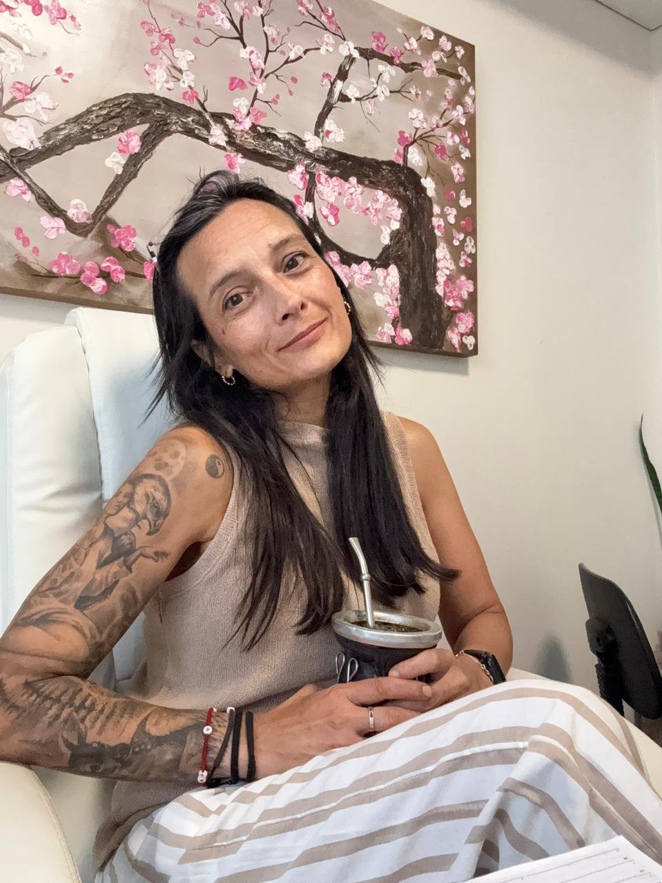 Mujer con tatuajes en el brazo izquierdo, sosteniendo una taza con popote, sentada en silla blanca, con fondo de pintura de árbol con flores rosadas.