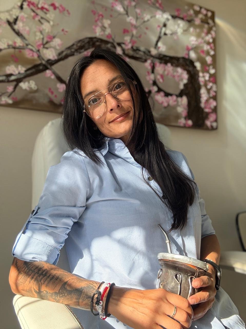 Mujer con gafas, camisa azul, y tatuajes en el brazo, sosteniendo un recipiente metálico, sentada frente a fondo con un cuadro de ramas de cerezo en flor.