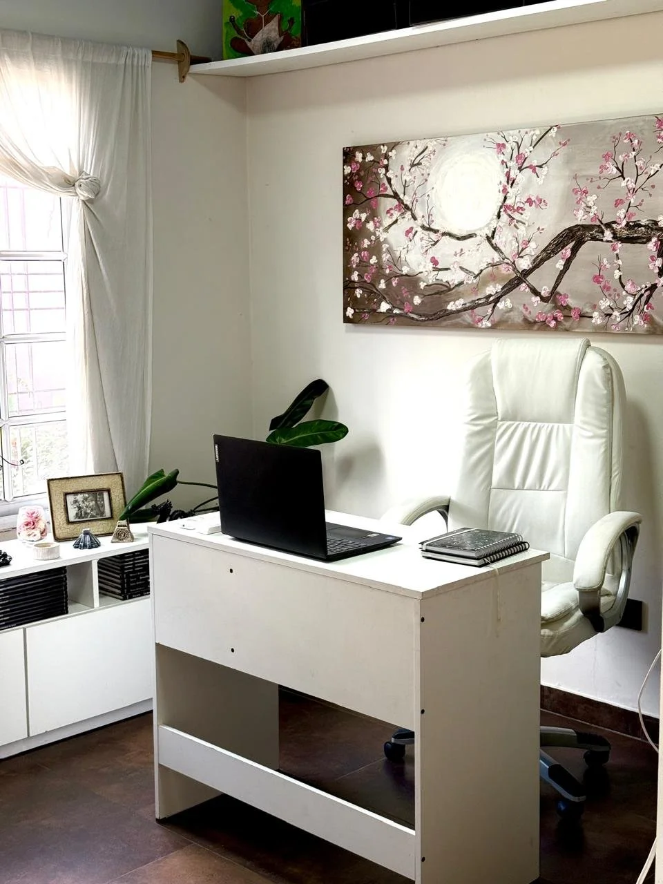 Oficina con silla blanca, laptop, libros, cuadro con flores de cerezo en la pared, ventana con cortinas blancas y una repisa con decoraciones.