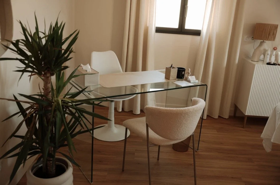 Une pièce élégante avec une table en verre, une chaise blanche et une chaise beige, un potted plant, une fenêtre avec des rideaux, une commode blanche avec une lampe et des objets décoratifs.