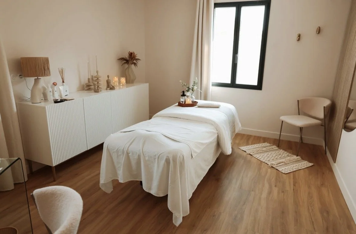 Une pièce lumineuse avec un lit à linge blanc, un fauteuil beige, une fenêtre et une commode blanche décorée de lampes, vases et décorations.