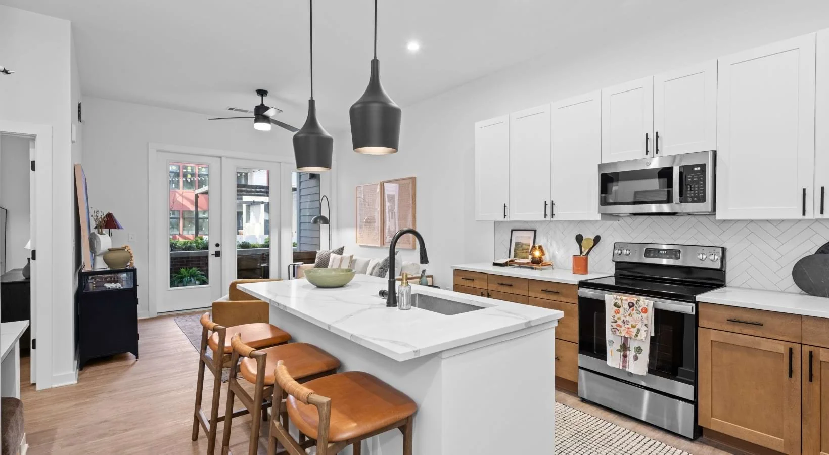 Kitchen with white upper cabinets, wood lower cabinets, stainless steel oven and microwave, white marble island with black sink and faucet, three black pendant lights, and a framed picture on the counter.