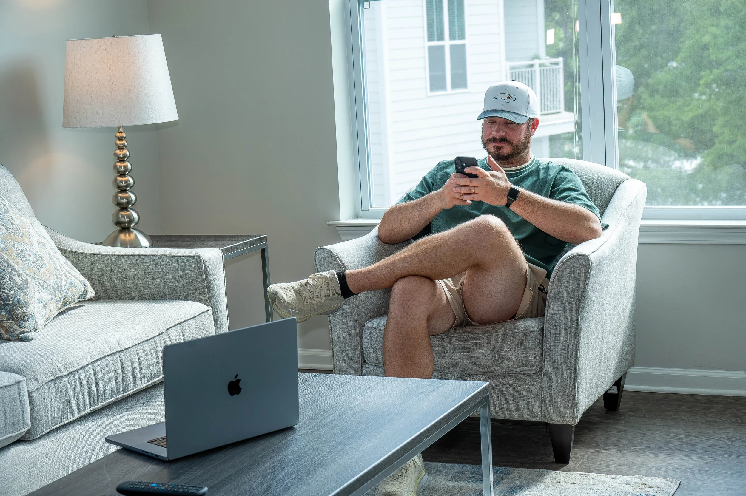 A man with a beard and mustache sitting on a white armchair, wearing a gray cap with a logo, green t-shirt, tan shorts, and white sneakers, looking at his phone, with a window showing trees and a neighboring house in the background, and a lamp on a table beside him.