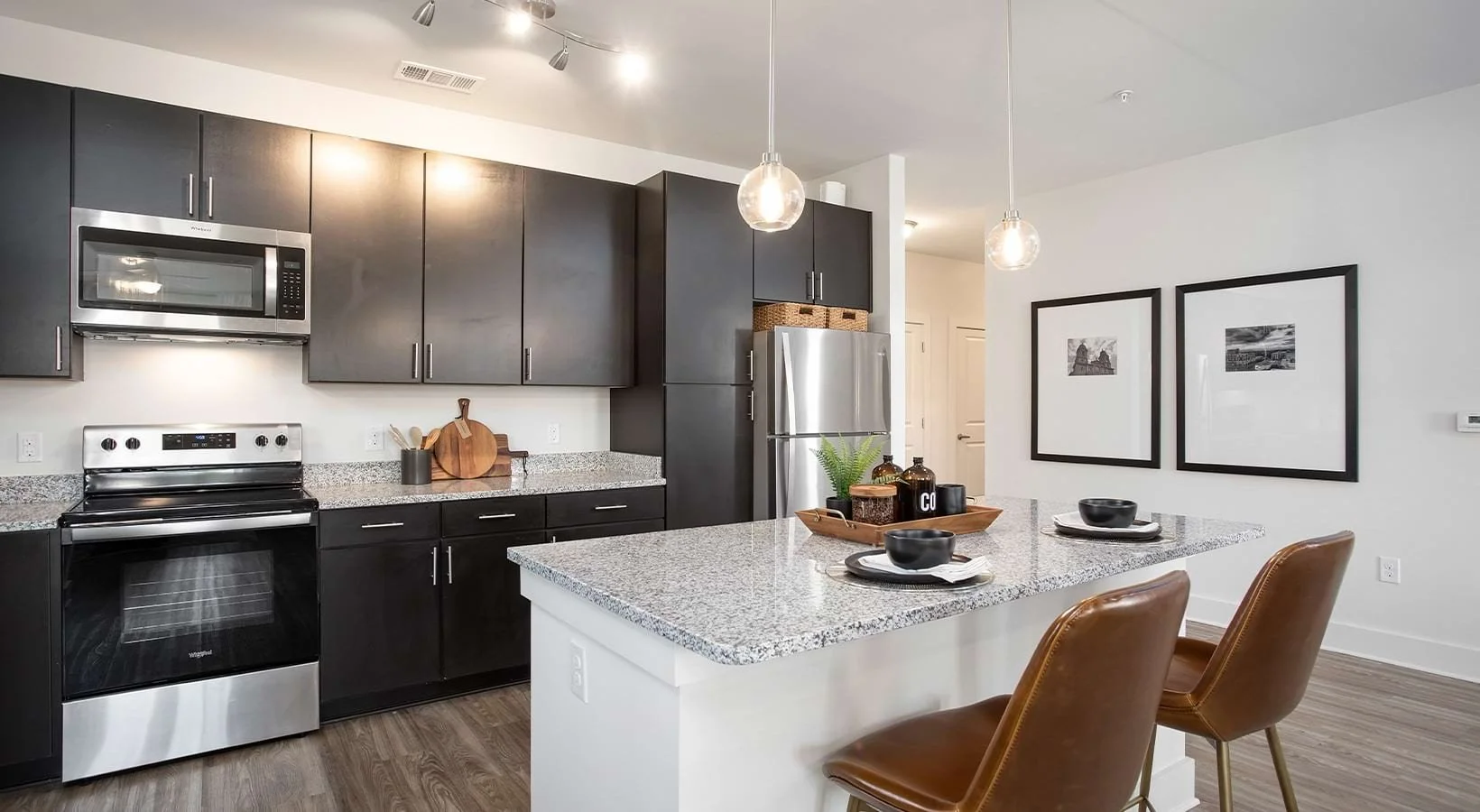 Modern kitchen with dark cabinets, stainless steel appliances, granite countertop island, and two brown leather chairs, decorated with black bowls, black and white framed wall art, and greenery.