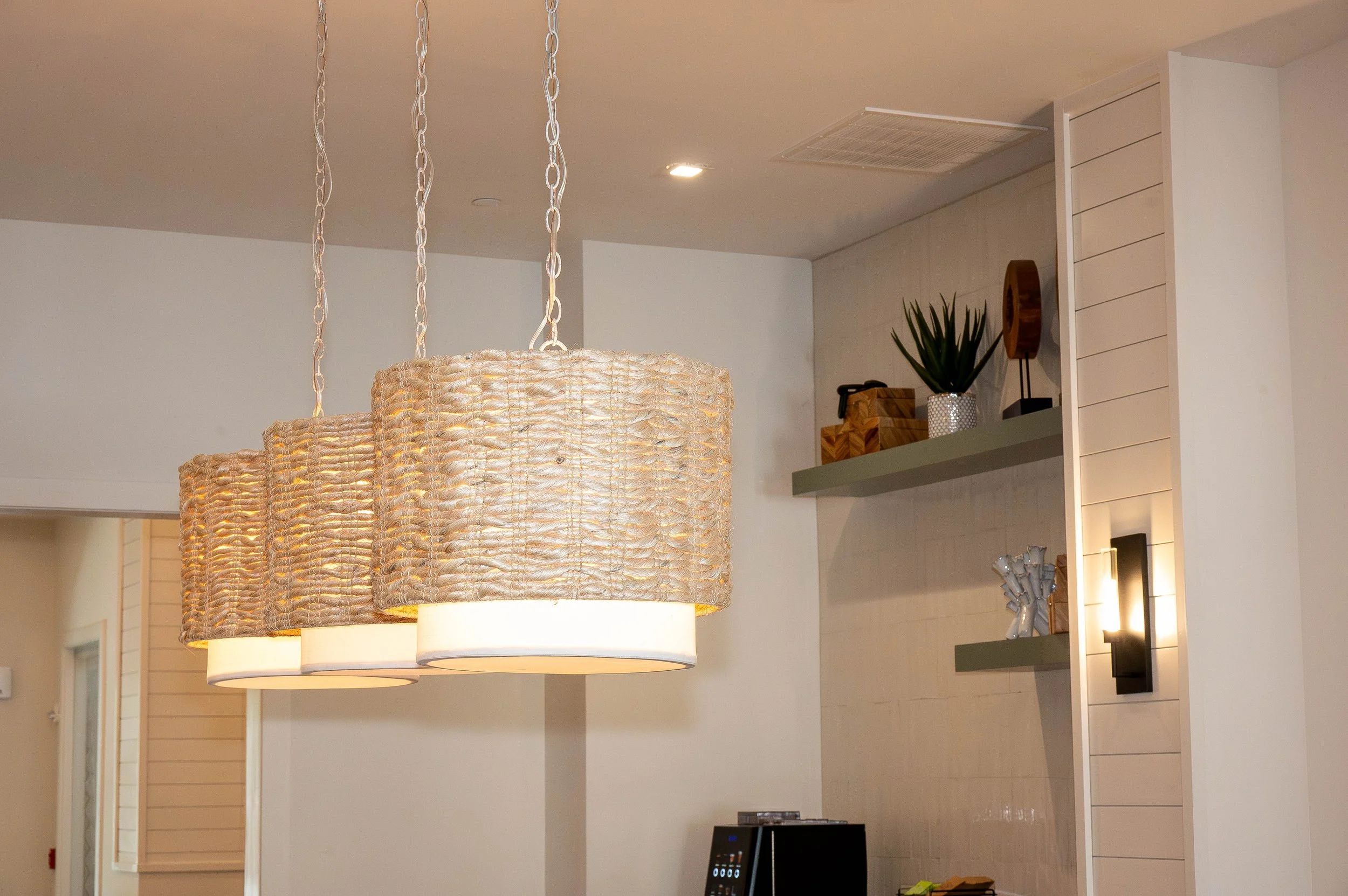 A modern interior space with hanging woven light fixtures, a shelf with decorative items including a potted plant, wooden boxes, and an abstract sculpture. The wall features horizontal paneling and a partially visible flat-screen TV.