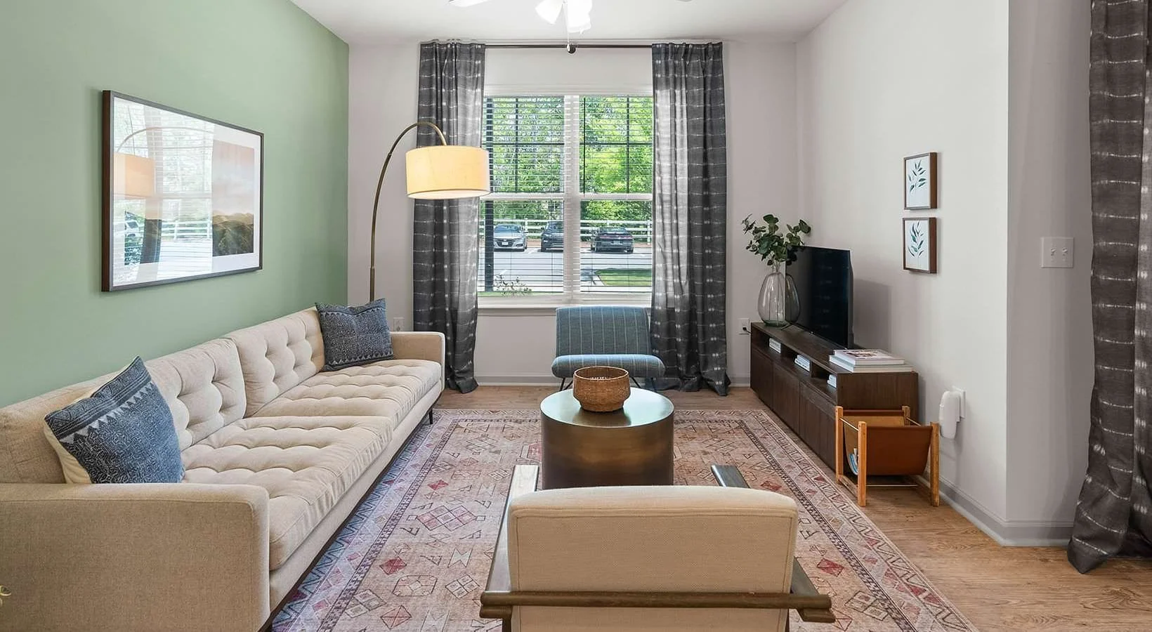 Living room with a beige tufted sofa, blue pillows, a floor lamp, a window with striped curtains, a chair, a TV on a wooden stand, artwork on the walls, and a patterned rug on wooden flooring.