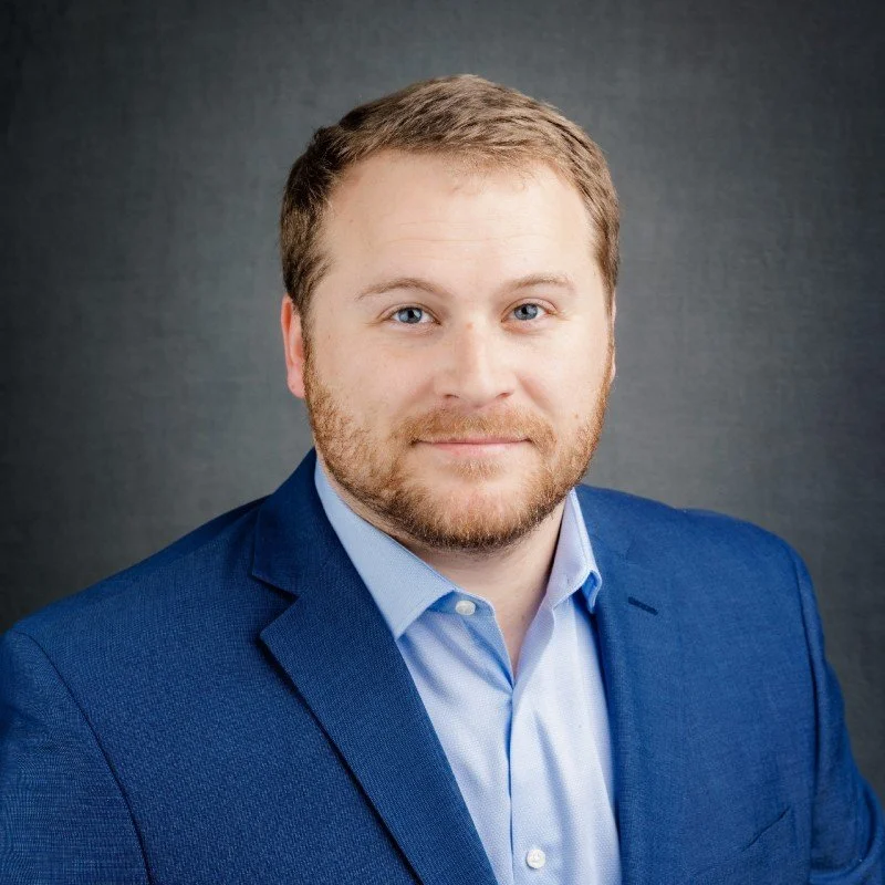 Headshot of a man with short light brown hair and a beard, wearing a blue blazer and light blue shirt, smiling against a dark gray background.