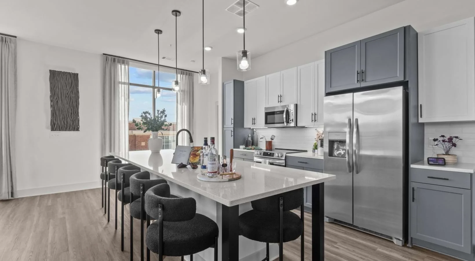 Modern kitchen with white and gray cabinetry, stainless steel appliances, a large island with black chairs, and a window with sheer white curtains showing a blue sky outside.