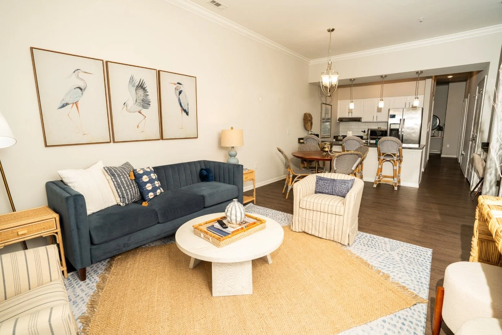 Living room with navy sofa, striped and patterned pillows, round white coffee table, beige armchair, artwork of three herons on the wall, wooden side table, light blue lamp, rug, open kitchen with white cabinetry, dining table with rattan chairs, pendant lighting.
