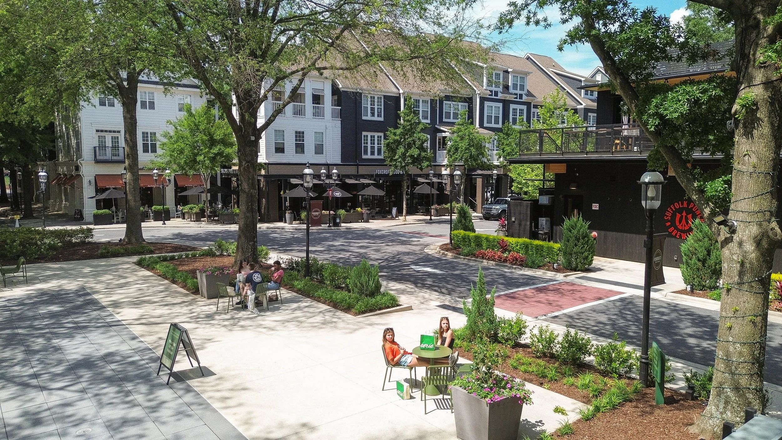 A lively outdoor shopping and dining area with trees, benches, and several people sitting and walking. Multi-story buildings with retail spaces and a brewery are visible in the background.