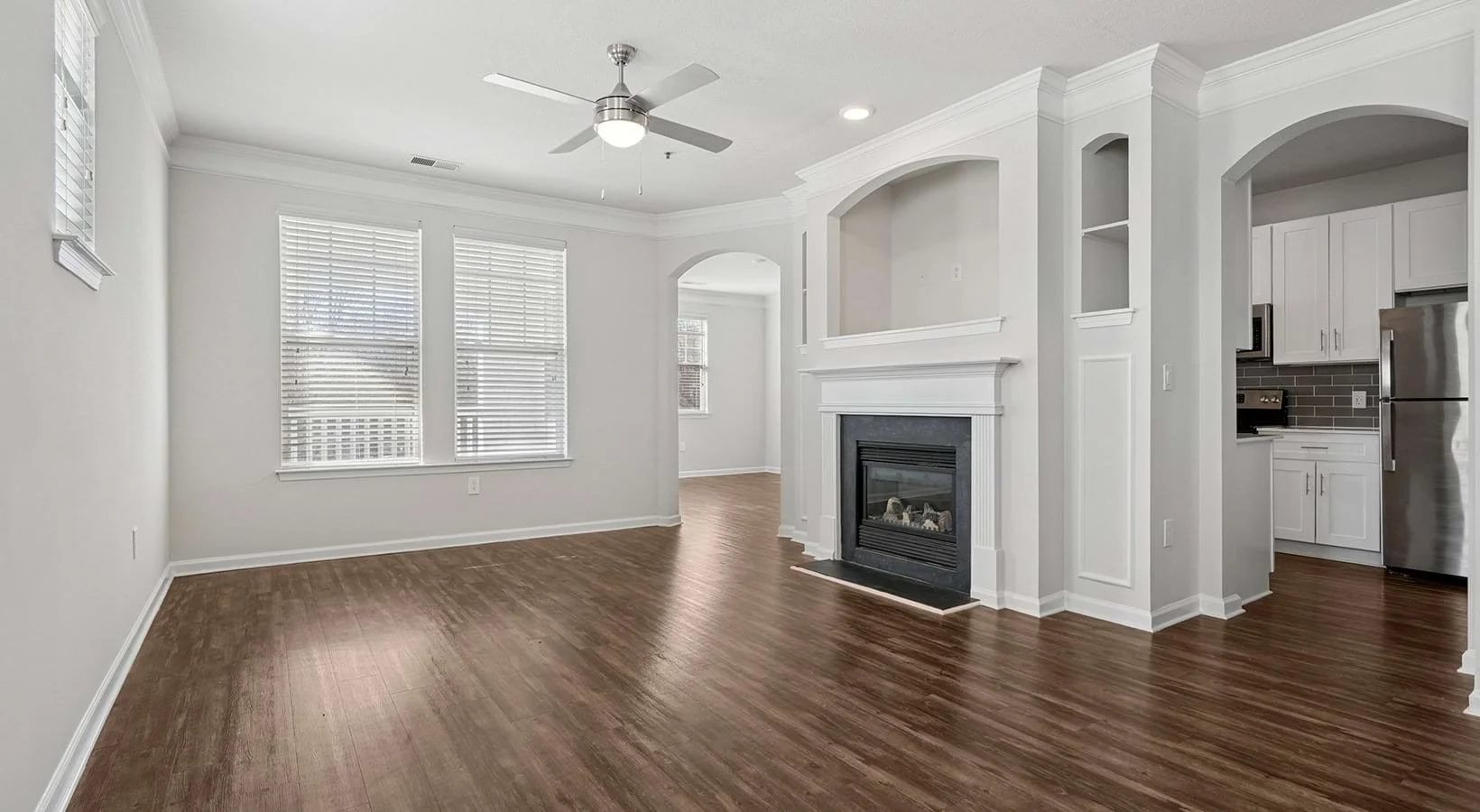 Spacious living room with hardwood floors, white walls, large windows with blinds, ceiling fan, and a fireplace with a white mantel. Kitchen area with white cabinets and stainless steel appliances visible in the background.