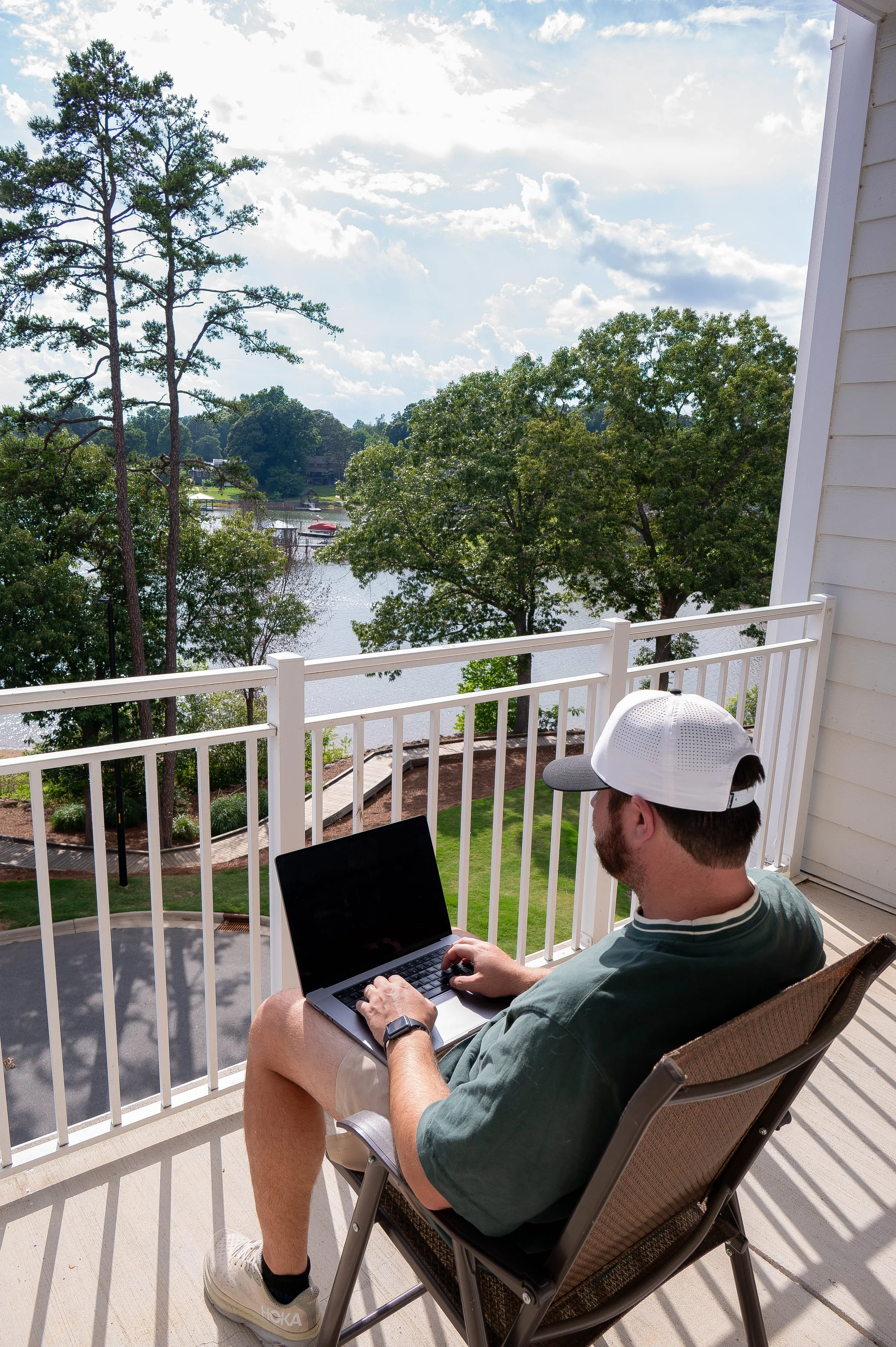 A man sitting on a balcony with a laptop, wearing a white cap, green shirt, beige shorts, and white sneakers, with a view of trees, water, and boats in the background under a partly cloudy sky.