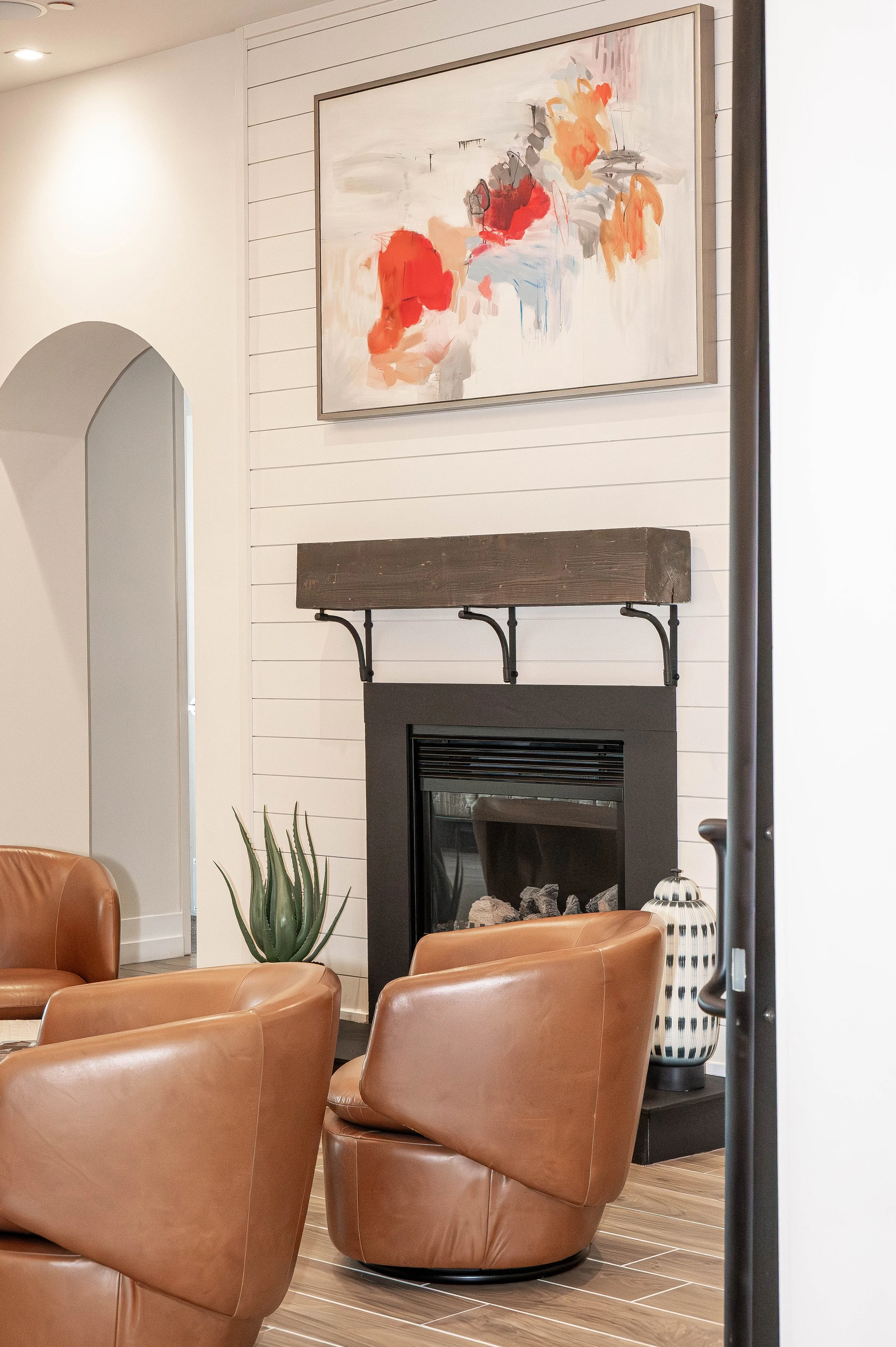 Interior of a modern living room featuring tan leather recliner chairs arranged around a fireplace with a wooden mantel, a potted agave plant, an abstract watercolor painting on the wall, and decorative vase near the fireplace.