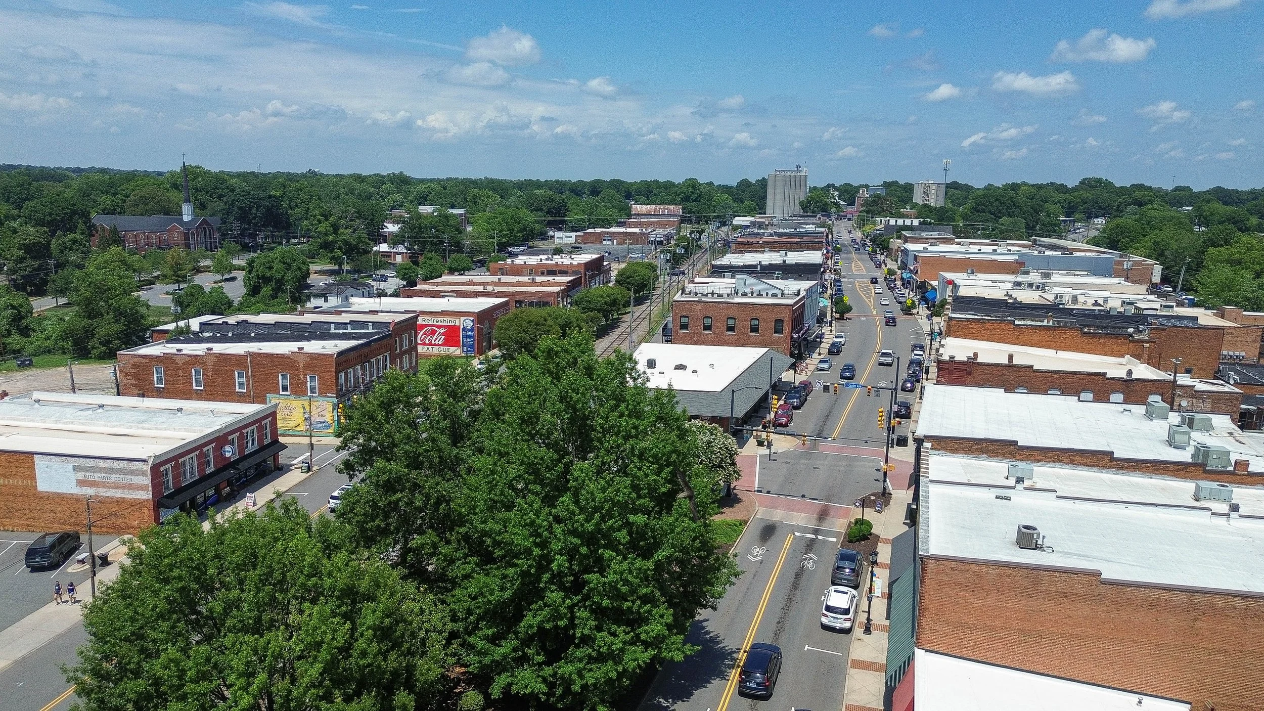 Aerial view of a small town with brick buildings, parked cars, a church with a tall steeple, and streets lined with trees on a sunny day.