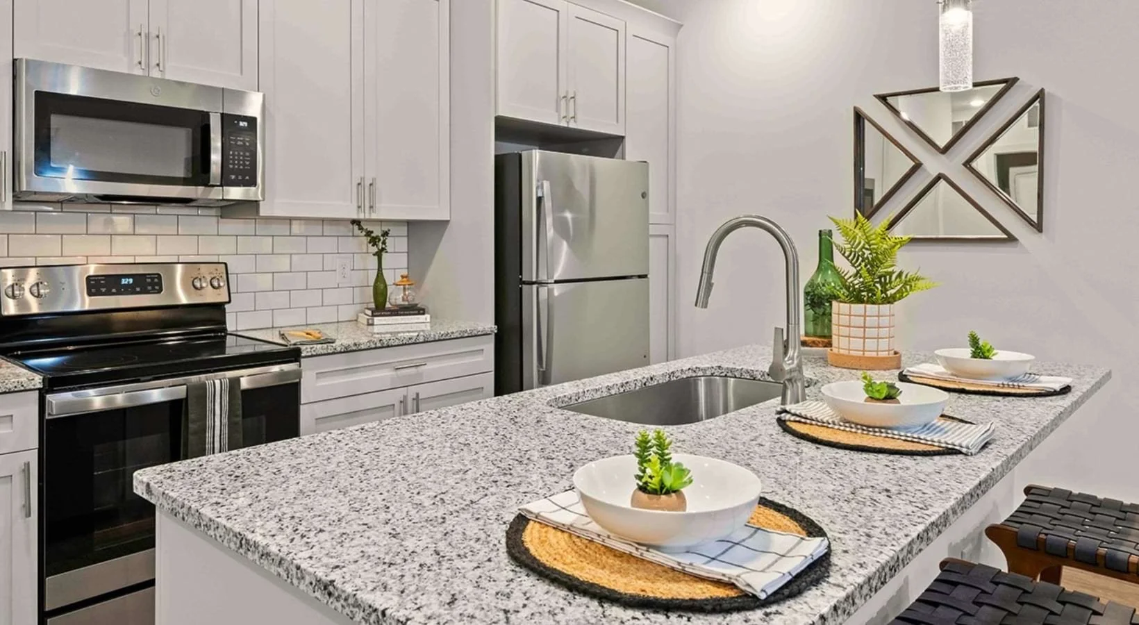 Modern kitchen with gray cabinets, stainless steel appliances, a granite countertop island, white subway tile backsplash, decorative items, and potted plants.
