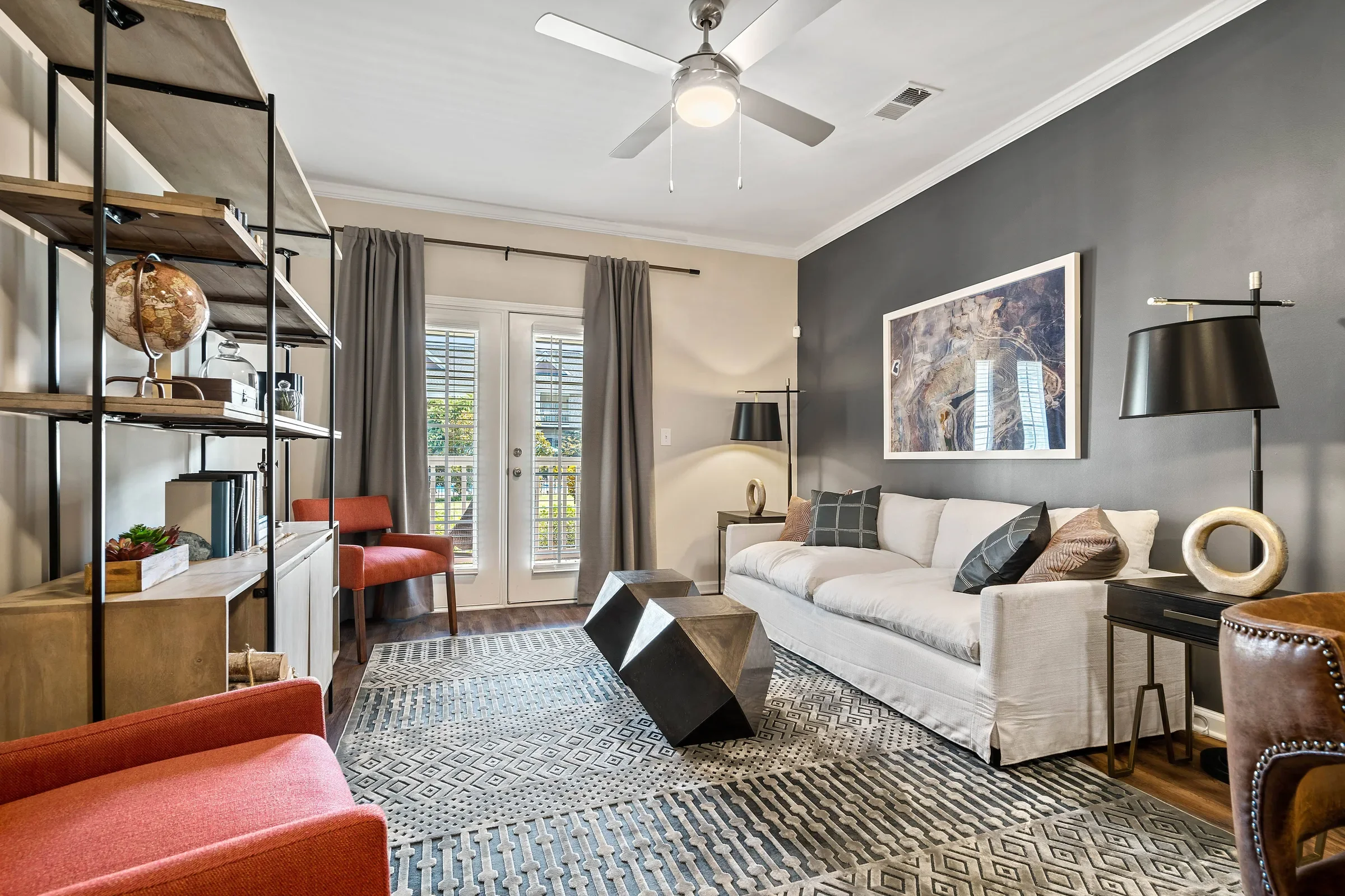 A living room featuring a white sofa with decorative pillows, modern black side lamps, a large artwork on a dark gray wall, a coffee table with geometric design, a patterned area rug, and a large window with blinds and gray curtains.
