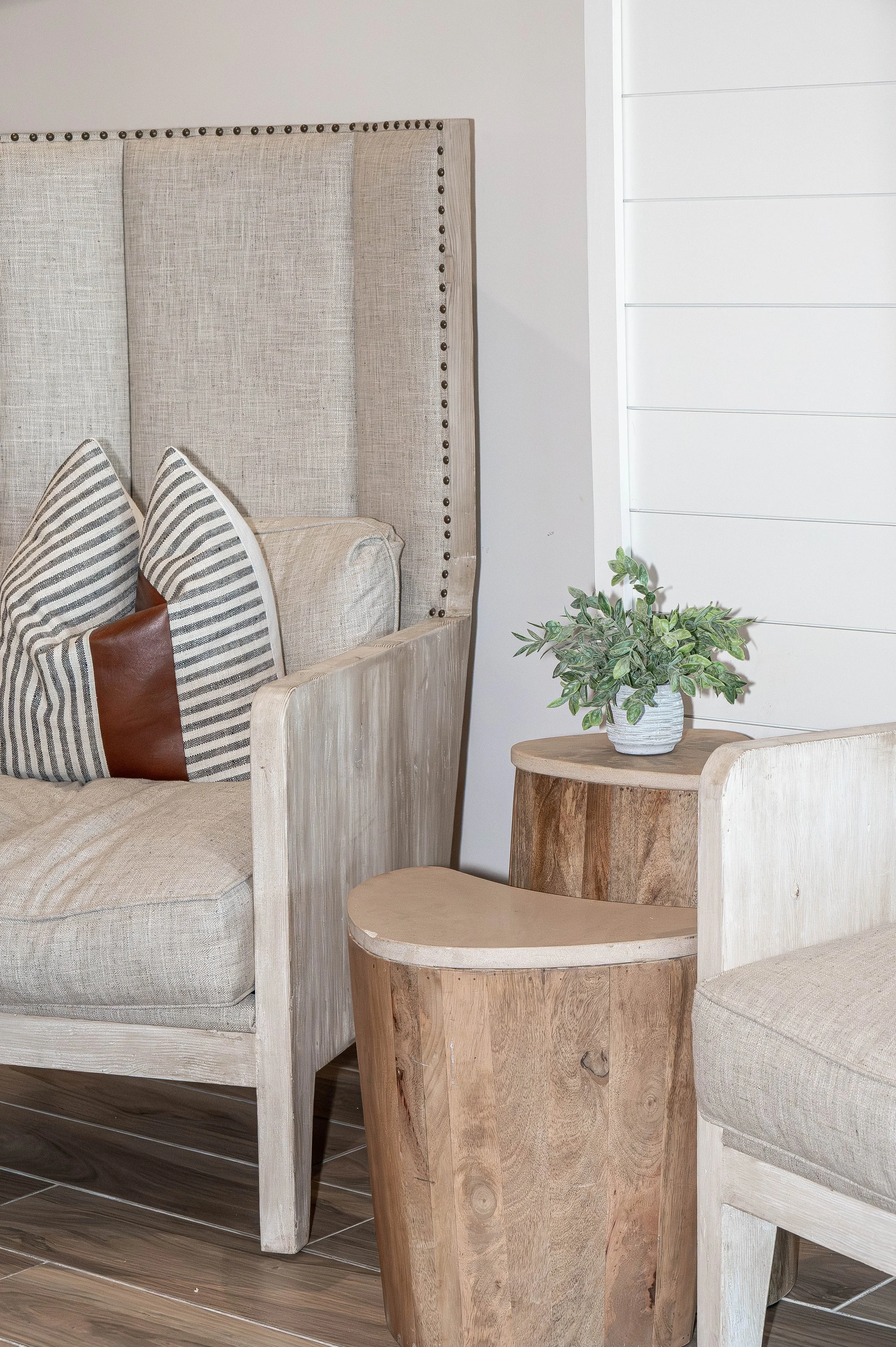 Living room corner with beige upholstered couch, striped and brown pillows, wood side table with potted green plant, light wood armchair, and white shiplap wall.