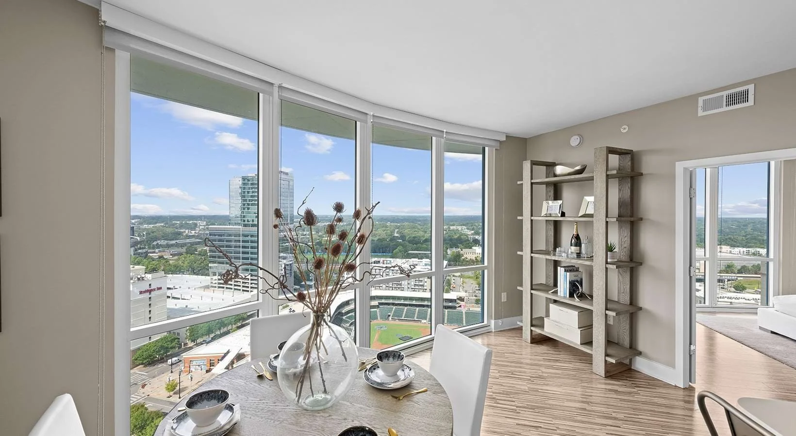 Modern apartment with large windows overlooking a cityscape, dining table with a glass vase and dishes, and a wooden bookshelf with decor.