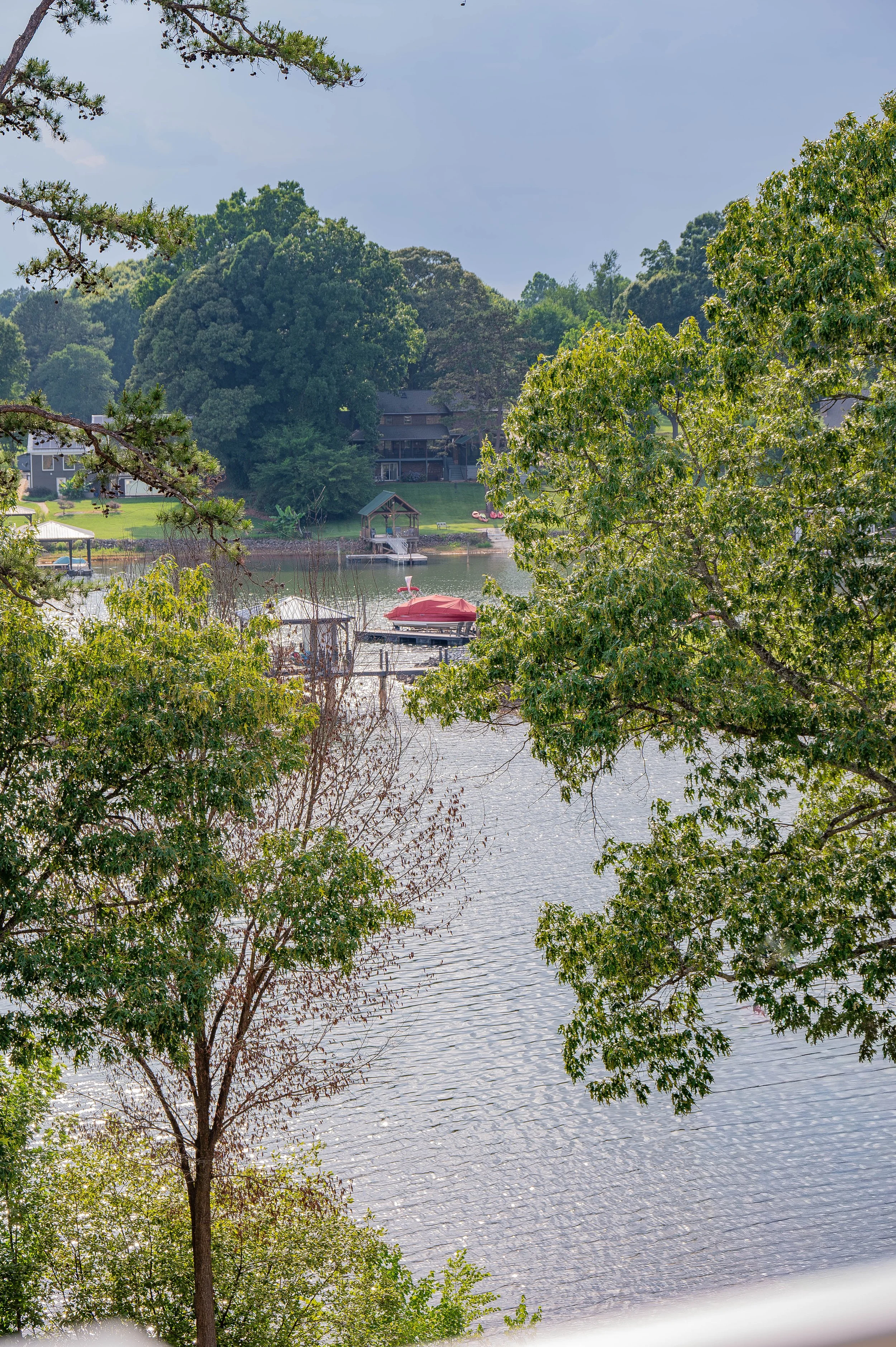 A scenic view of a lakeside landscape with trees in the foreground, and houses, a dock, and a boat with a red cover along the water in the background.
