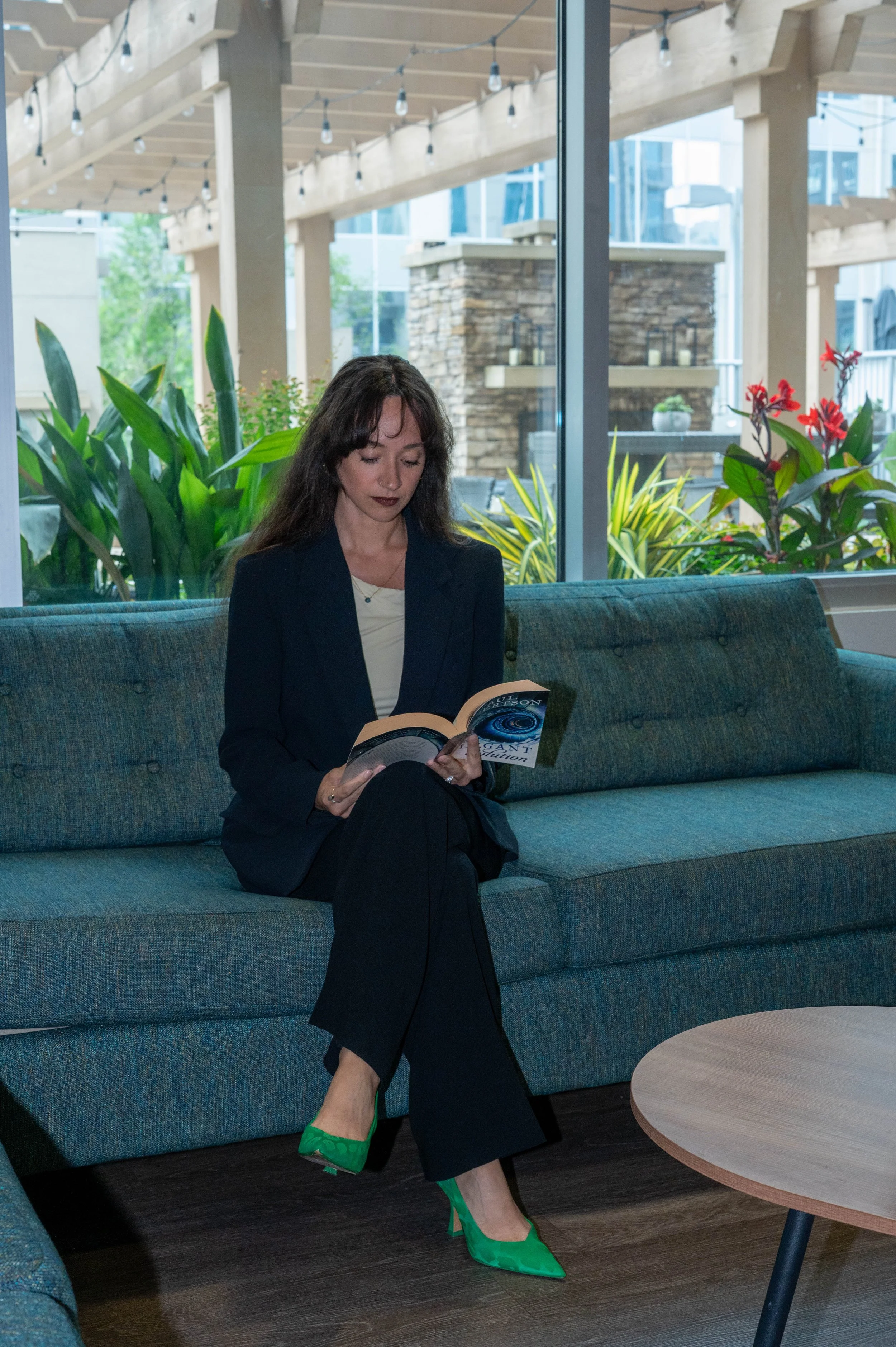 A woman with dark hair, wearing a black blazer and matching pants, sitting on a blue-green couch, reading a book with a cosmic cover. She wears green high heels and has dark lipstick. Behind her, there are green plants and large glass windows with a view of a patio with string lights and a stone wall.