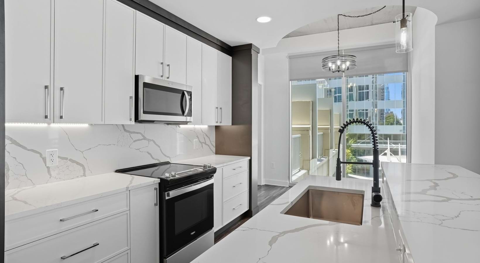 Modern kitchen with white cabinets, marble countertops, and a view of a balcony with glass doors, featuring a black faucet, stainless steel microwave, and stove.