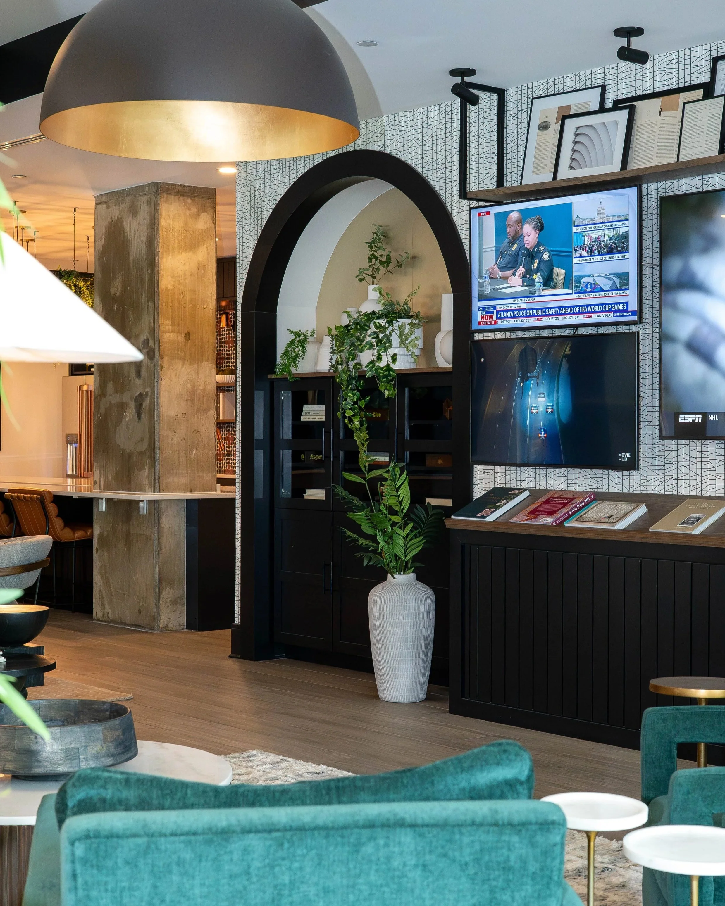 Interior of a modern living space with green velvet chairs, a black bookshelf, a large decorative white vase with green plants, two wall-mounted TVs displaying news, and a large hanging lamp with a black exterior and gold interior.