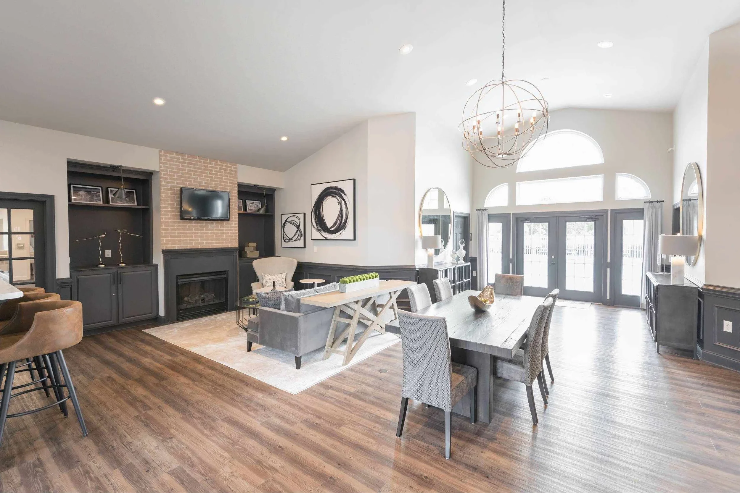 Bright living room with high ceiling, large windows, a chandelier, a fireplace with a TV above it, and a variety of chairs and tables.