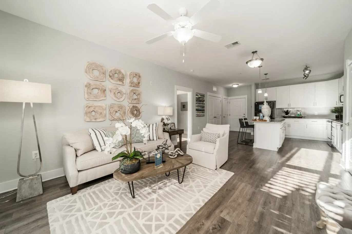 Living room with beige sofa, white armchair, wooden coffee table, decorative wall art, potted orchids, and hardwood floors, leading into a kitchen with white cabinets and a breakfast bar.