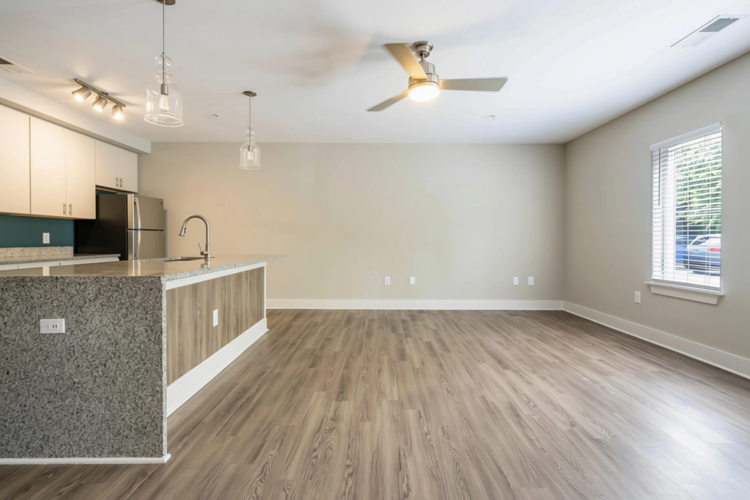 An open-concept living space with a kitchen island, beige walls, wooden flooring, large window with blinds, ceiling fan, and ceiling lights.