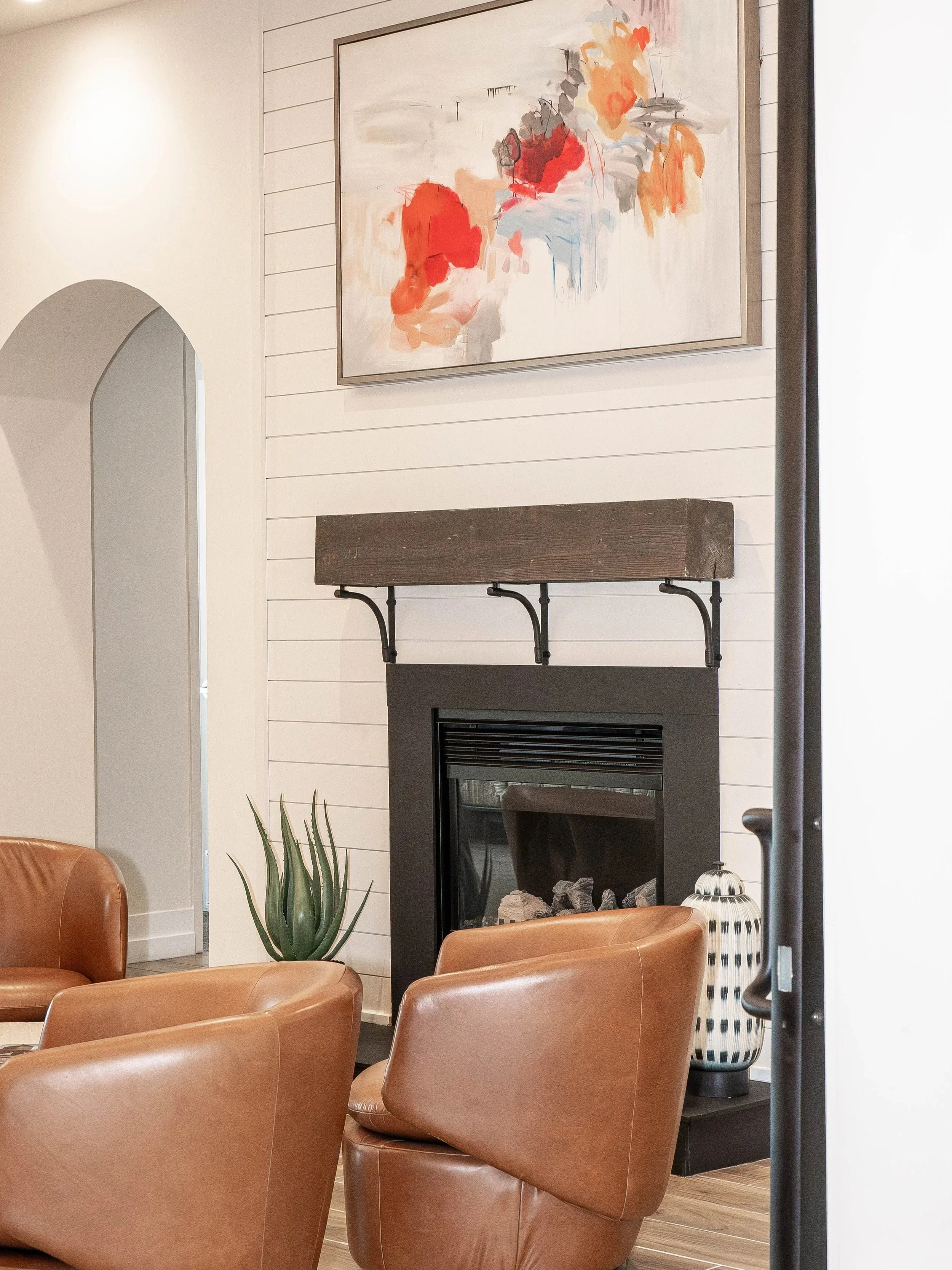Living room with leather chairs, a fireplace with a wooden mantel, a large abstract painting with red, orange, gray, and blue colors on the wall, a tall green plant in a pot, and a decorative vase near the fireplace.
