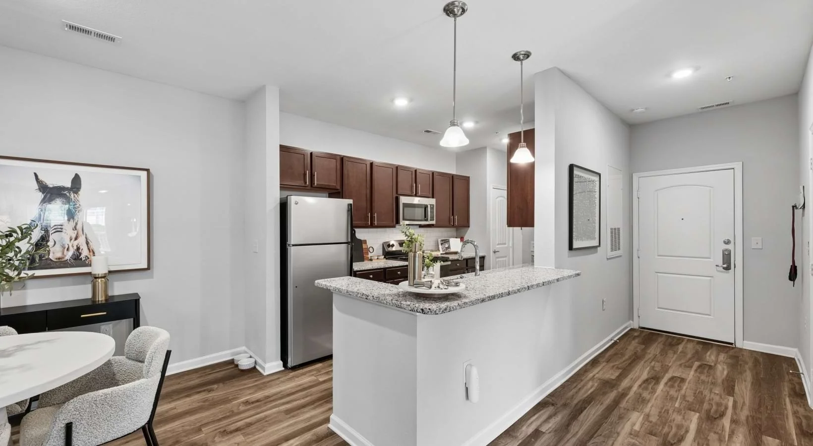 Modern kitchen with white walls, wooden cabinets, stainless steel refrigerator, microwave, and granite countertop. Adjacent dining area with a round table, four chairs, and a framed zebra picture on the wall.