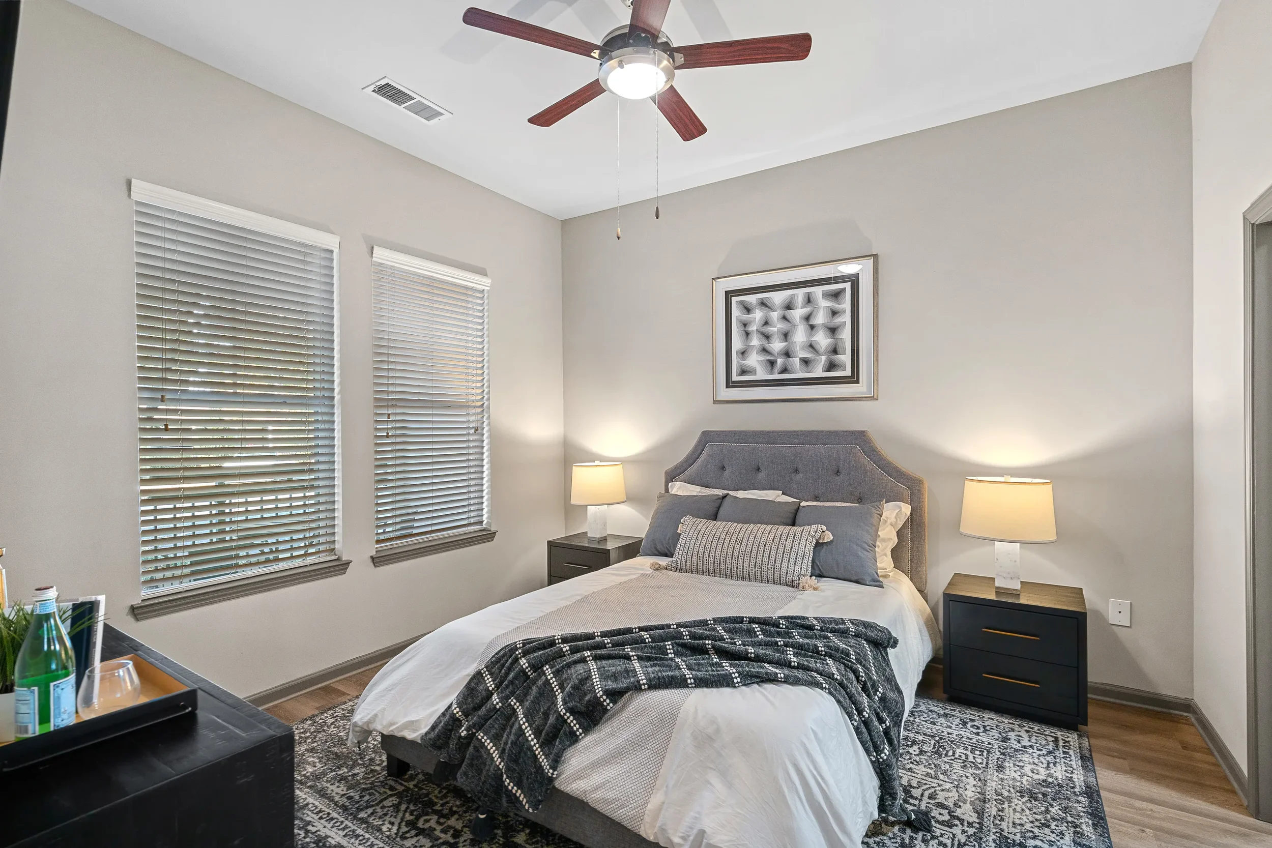 A bedroom with two windows covered with white blinds, a gray upholstered headboard, a bed with white and gray bedding, nightstands with lamps, a patterned rug, modern wall art, and a ceiling fan with wooden blades.