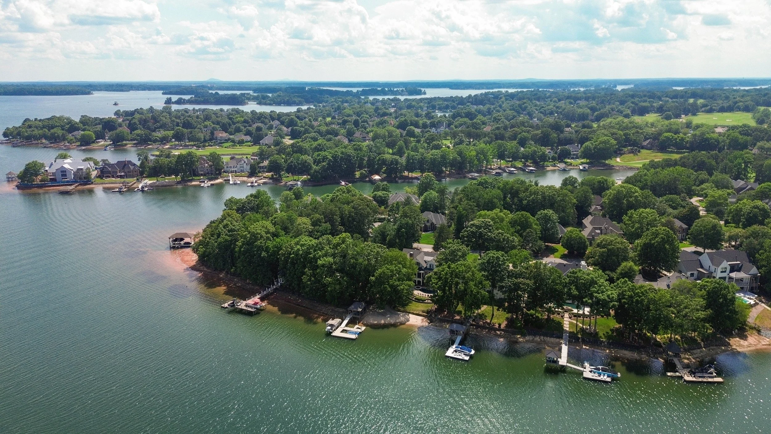 Aerial view of a lakeside residential neighborhood with houses, docks, and boats on the water, surrounded by lush green trees and open land, under a partly cloudy sky.