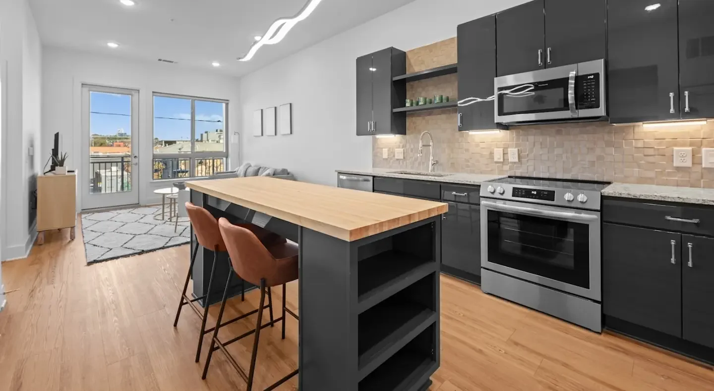 Modern kitchen with black cabinets, stainless steel appliances, a wooden countertop island, and a living area with large windows and a balcony door.