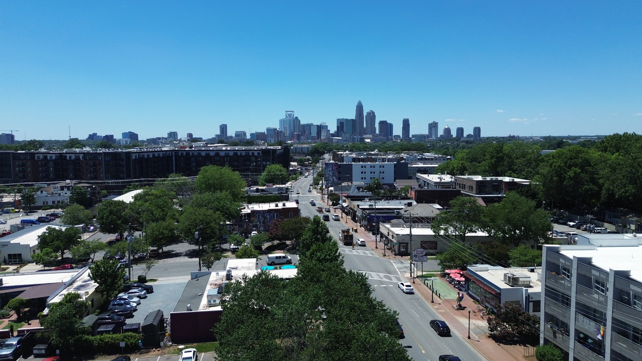 A city skyline with tall buildings and a clear blue sky, viewed from a suburban area with smaller buildings, trees, and streets in the foreground.