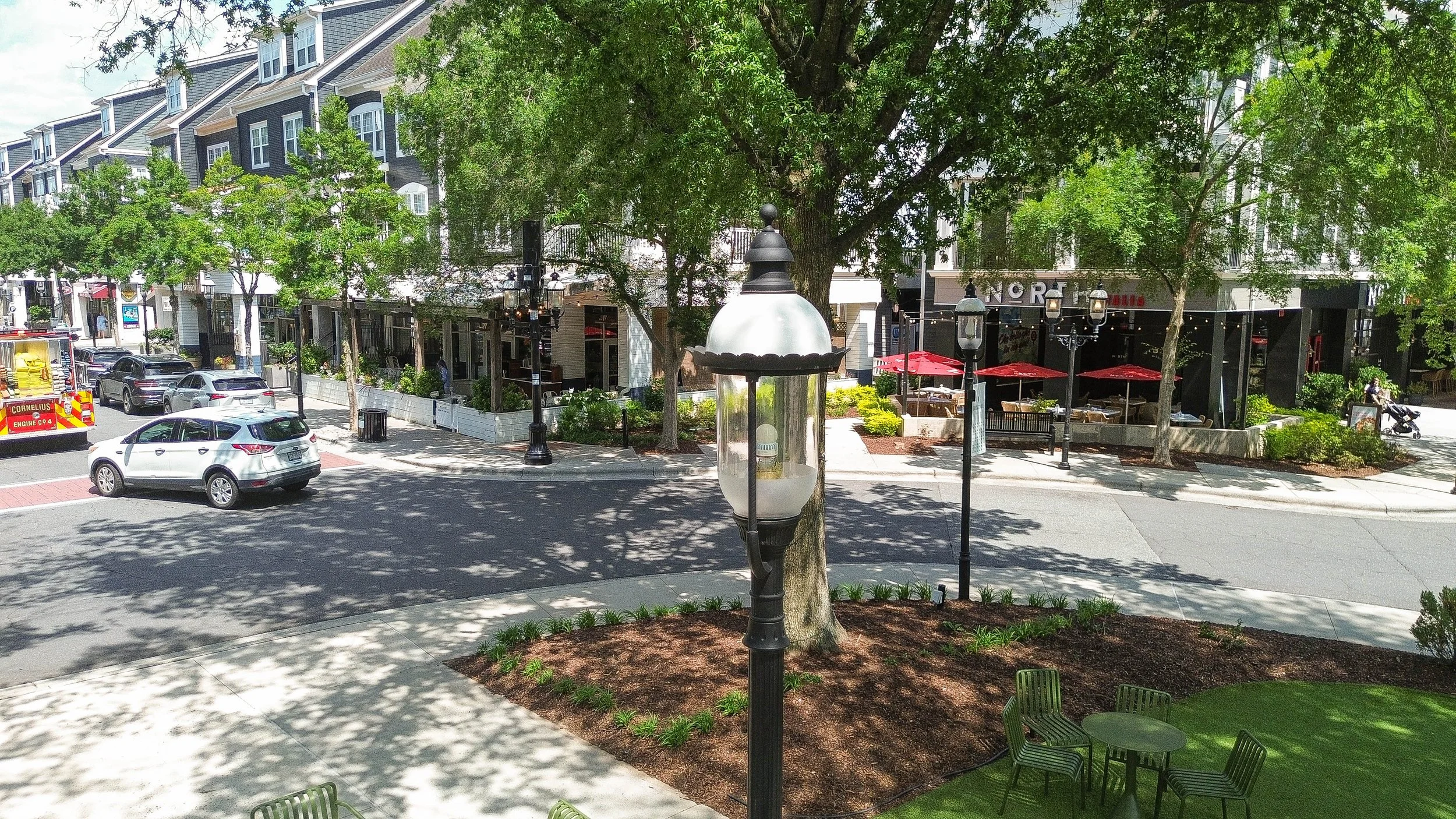 Street view showing parked cars, a fire truck, trees, outdoor seating area with red umbrellas, and a large building with storefronts. A decorative street lamp is in the foreground.